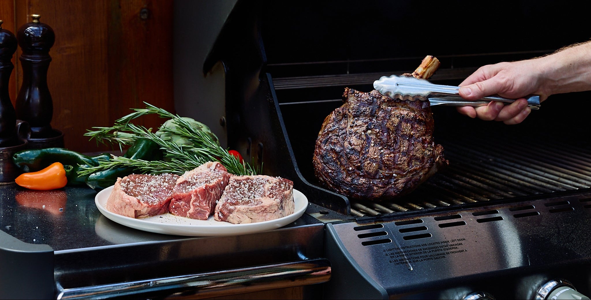 Person grilling a large piece of meat next to raw steaks on a plate with vegetables.